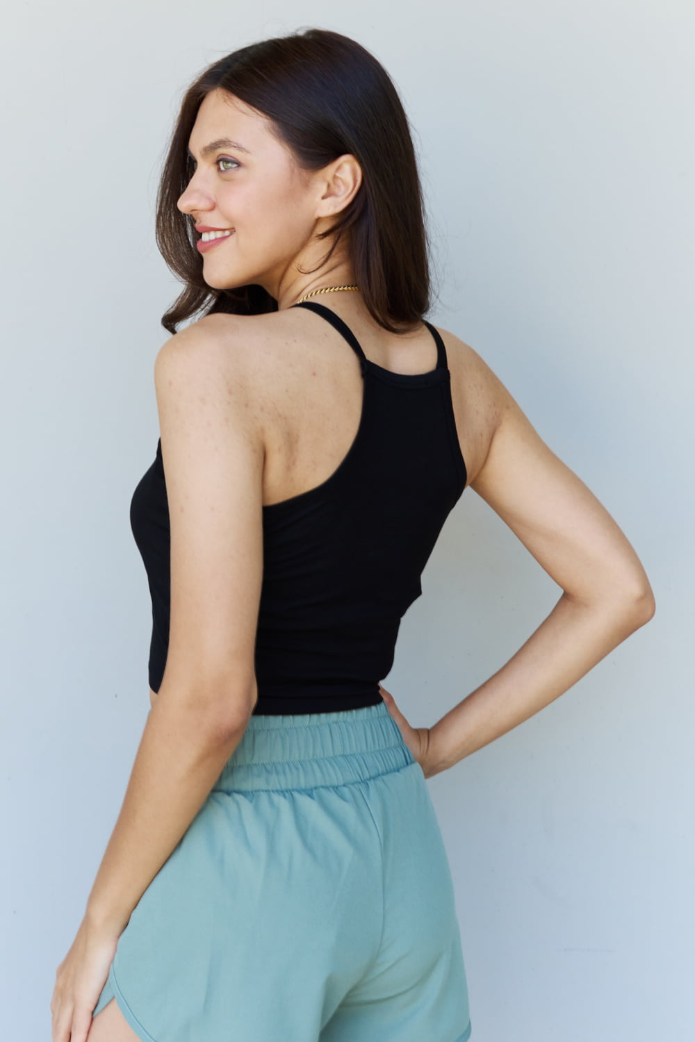 A person wearing a Ninexis Everyday Staple Soft Modal Short Strap Ribbed Tank Top in Black and light blue shorts is standing against a plain background, smiling slightly and looking to the side.