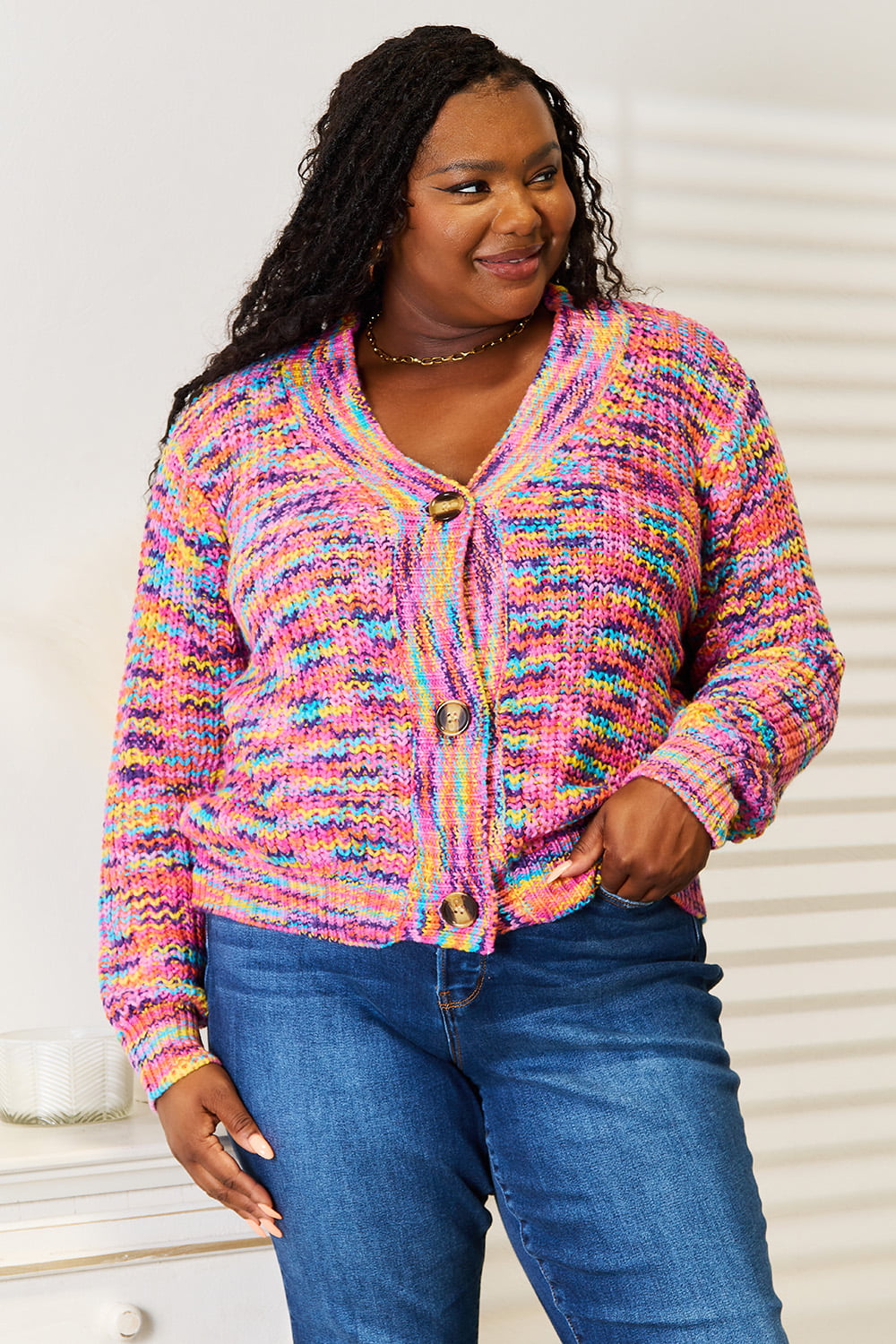 Person wearing an Angel Wings Woven Right V-Neck Long Sleeve Cardigan, characterized by its multicolored design and large buttons, paired with blue jeans. The cardigan, made of 100% acrylic knit, offers a versatile look perfect for any occasion. They are standing indoors against a white background.