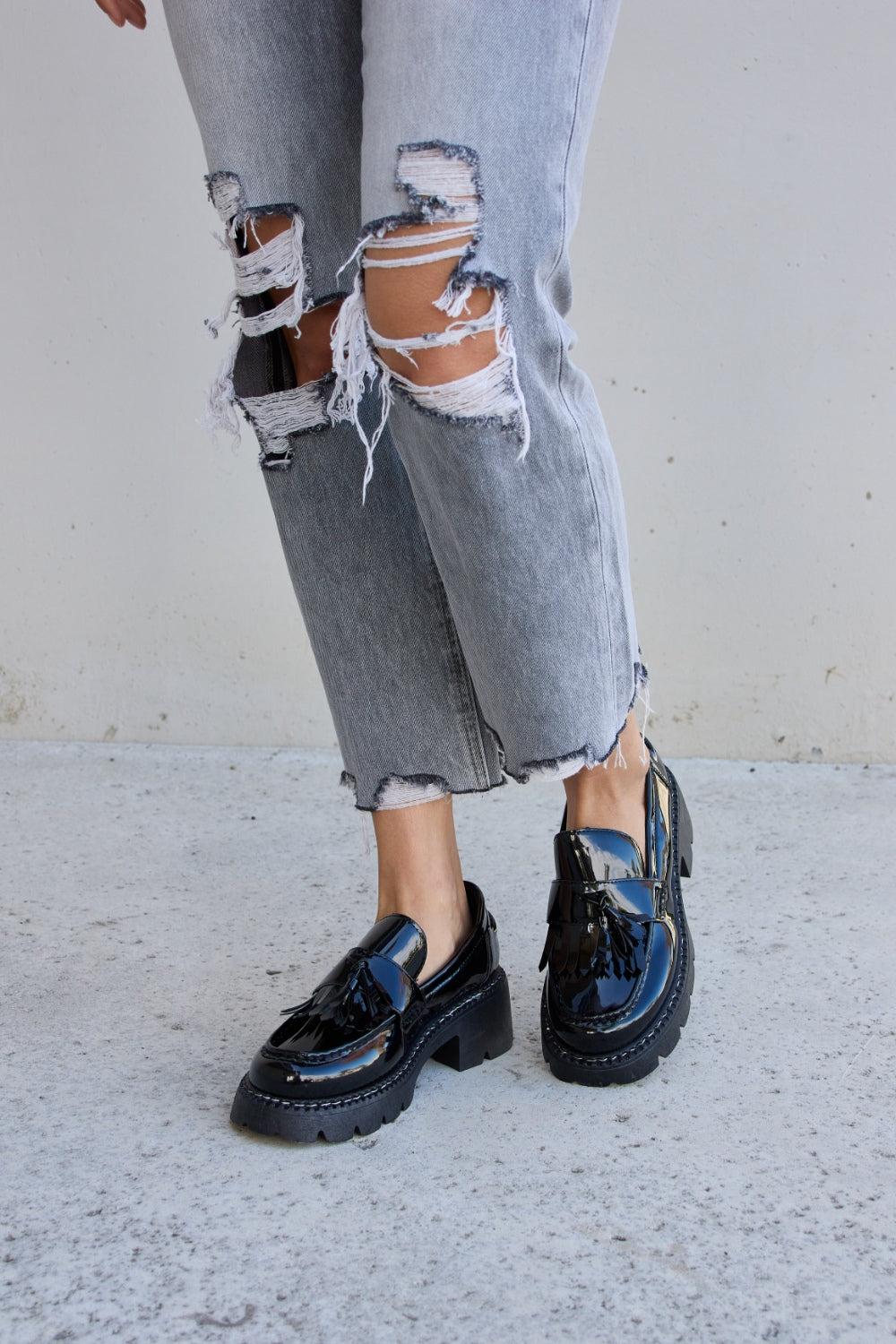 A person wearing ripped gray jeans and the Forever Link Tassel Detail Mid Heel Chunky Loafers, which feature shiny black tones, chunky soles, and a subtle tassel detail.