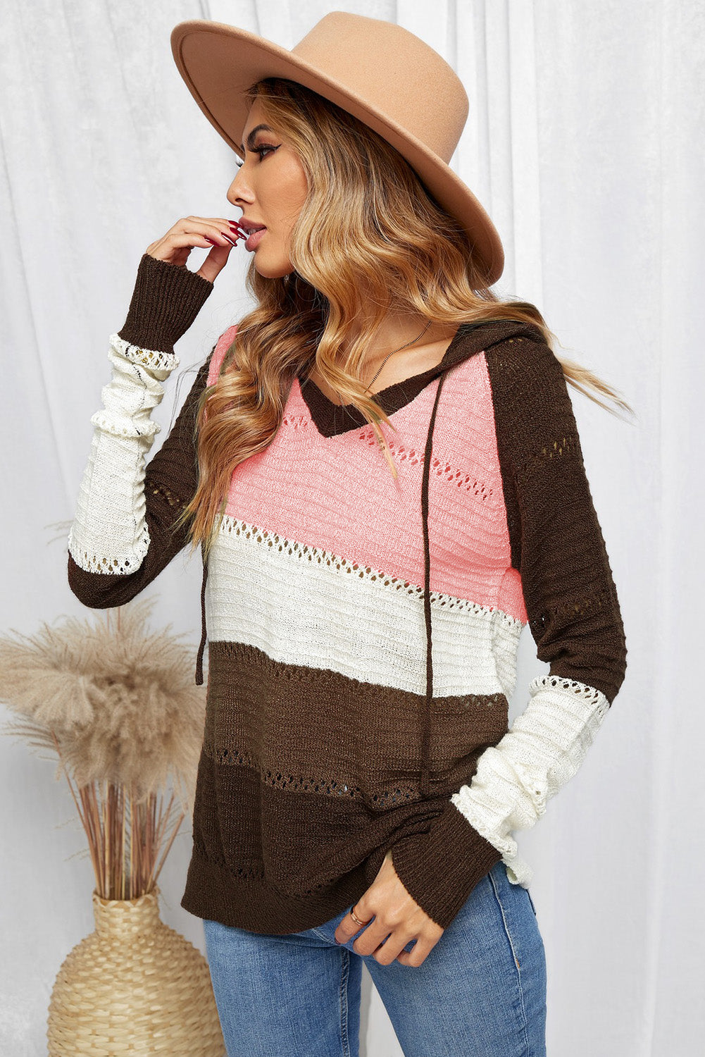 A person stands with their back to the camera, adjusting the hood of a Brown Beach Bonfire Knitted Hoodie featuring dark brown, pink, white, and light brown stripes. Perfect for a casual style, they are wearing distressed blue jeans and showcasing the laid-back vibe of lightweight hoodies.