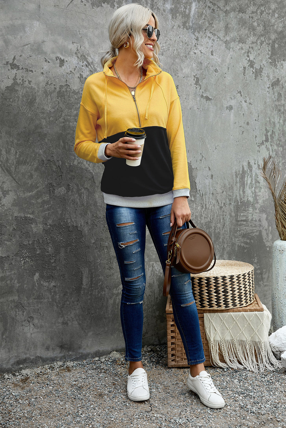 A person with long, light hair in a ponytail, wearing a Black Zipped Colorblock Sweatshirt with Pockets, blue jeans, and glasses, is holding a white cup and a brown handbag, shown from behind against a gray wall.