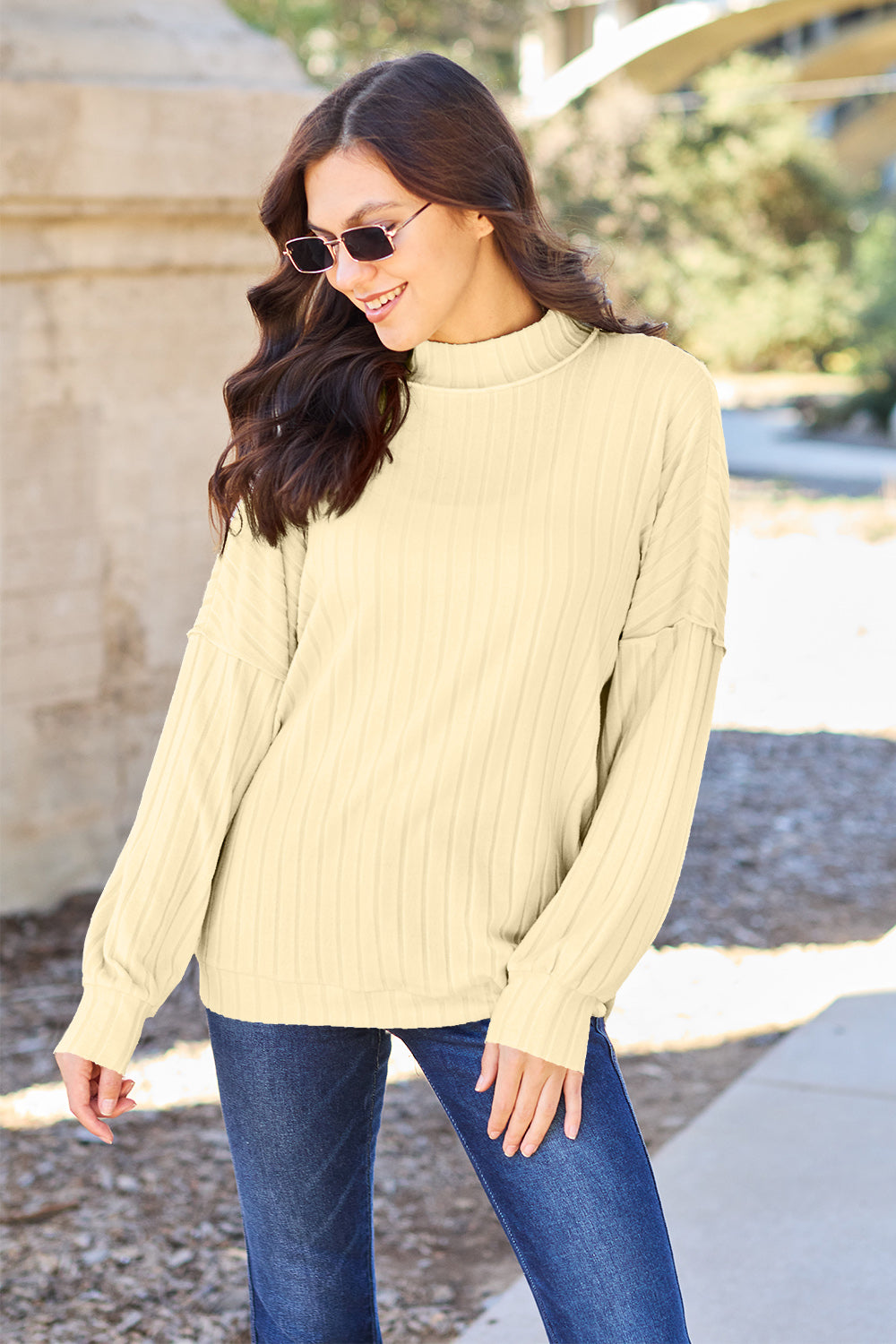 A woman with long hair is wearing a green Basic Bae Full Size Ribbed Exposed Seam Mock Neck Knit Top, blue jeans, and sunglasses. She is standing outdoors and smiling.