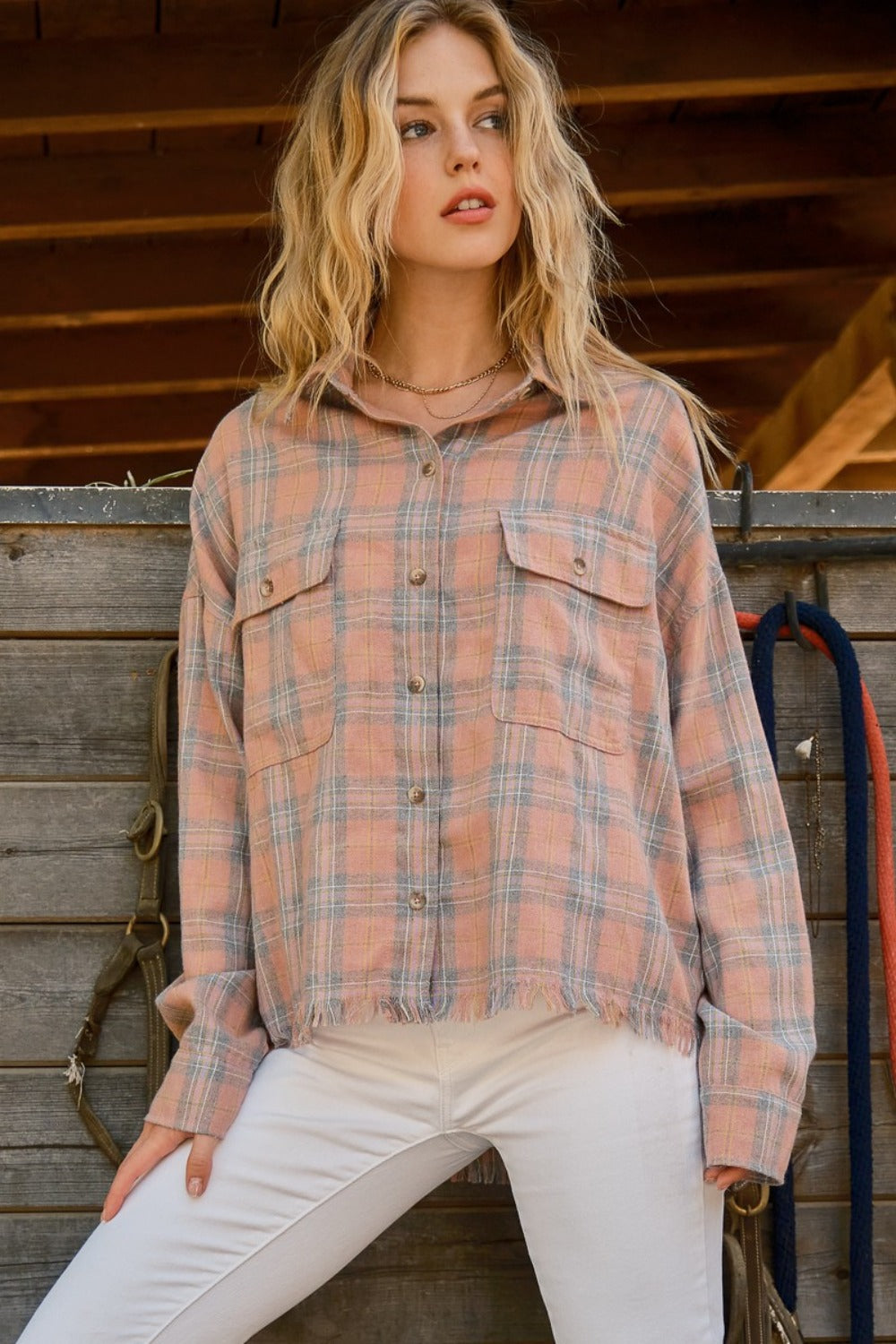 A person wearing the And The Why Full Size Plaid Button Up Raw Hem Shirt and white pants is standing in front of a wooden structure, showcasing raw edge detailing, with bridles hanging beside them.