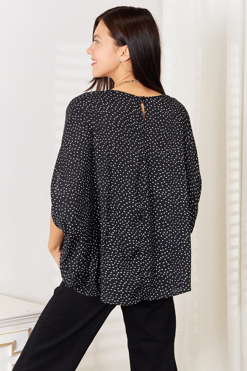 A woman stands wearing the Perfee Printed Dolman Sleeve Round Neck Blouse with white dots and black pants, smiling and looking to her left. The blouse features a printed design that adds a touch of elegance to her outfit.