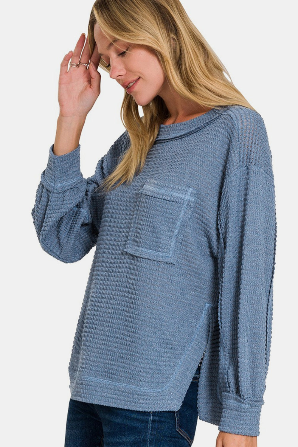 A woman wearing the Zenana Exposed Seam Side Slit Long Sleeve Top, which features a loose fit and textured blue fabric with side slits and a pocket on the left side, stands against a plain, light background. She has blonde hair and is looking directly at the camera.