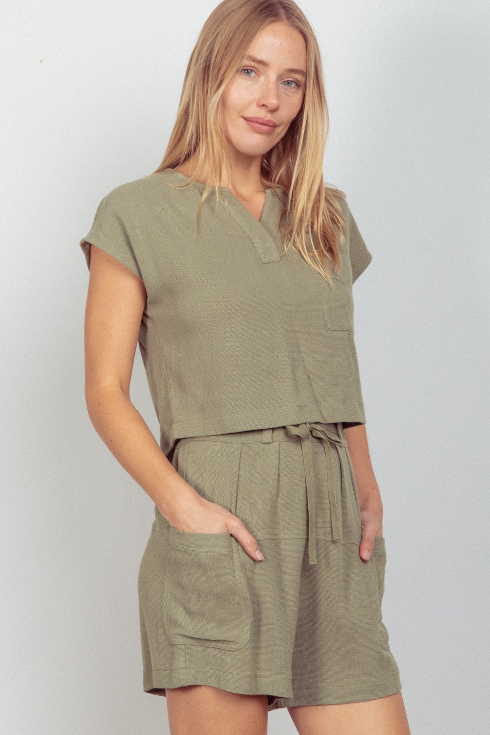 A person wearing the VERY J Woven Cropped Top & Waist Tie Shorts Set in khaki green, which includes a short-sleeved cropped top and matching waist tie shorts crafted from woven fabric, stands against a plain background.