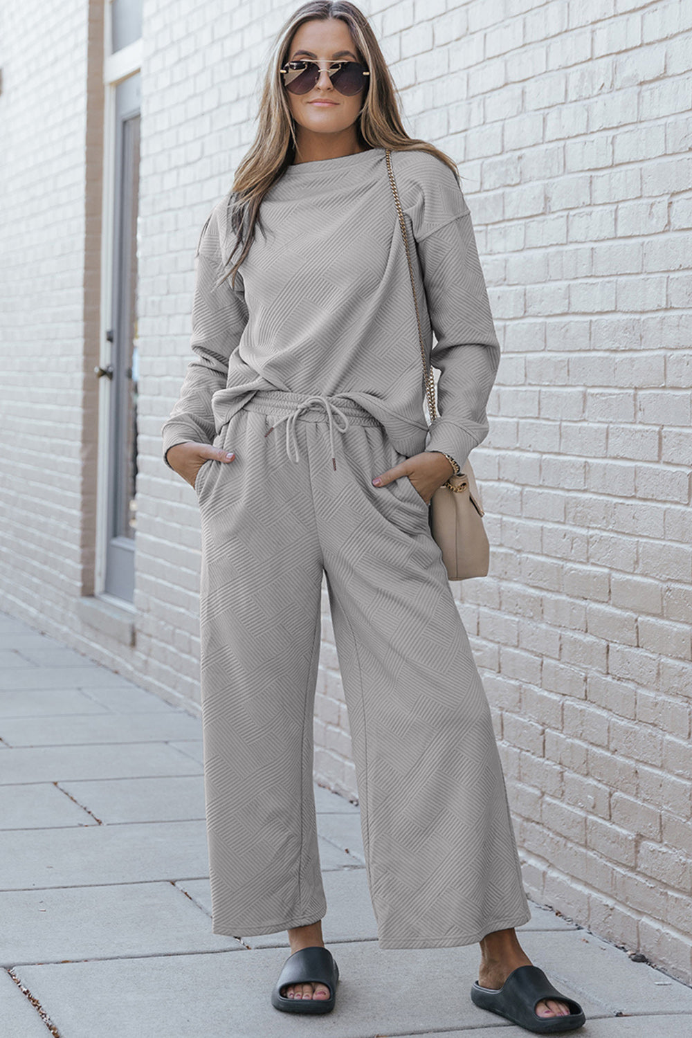 A woman with blonde hair, dressed in the Double Take Full Size Textured Long Sleeve Top and Drawstring Pants Set featuring a stretchy black long-sleeve top and matching wide-leg trousers made of rayon spandex, stands barefoot in a room with white walls and a wooden floor.