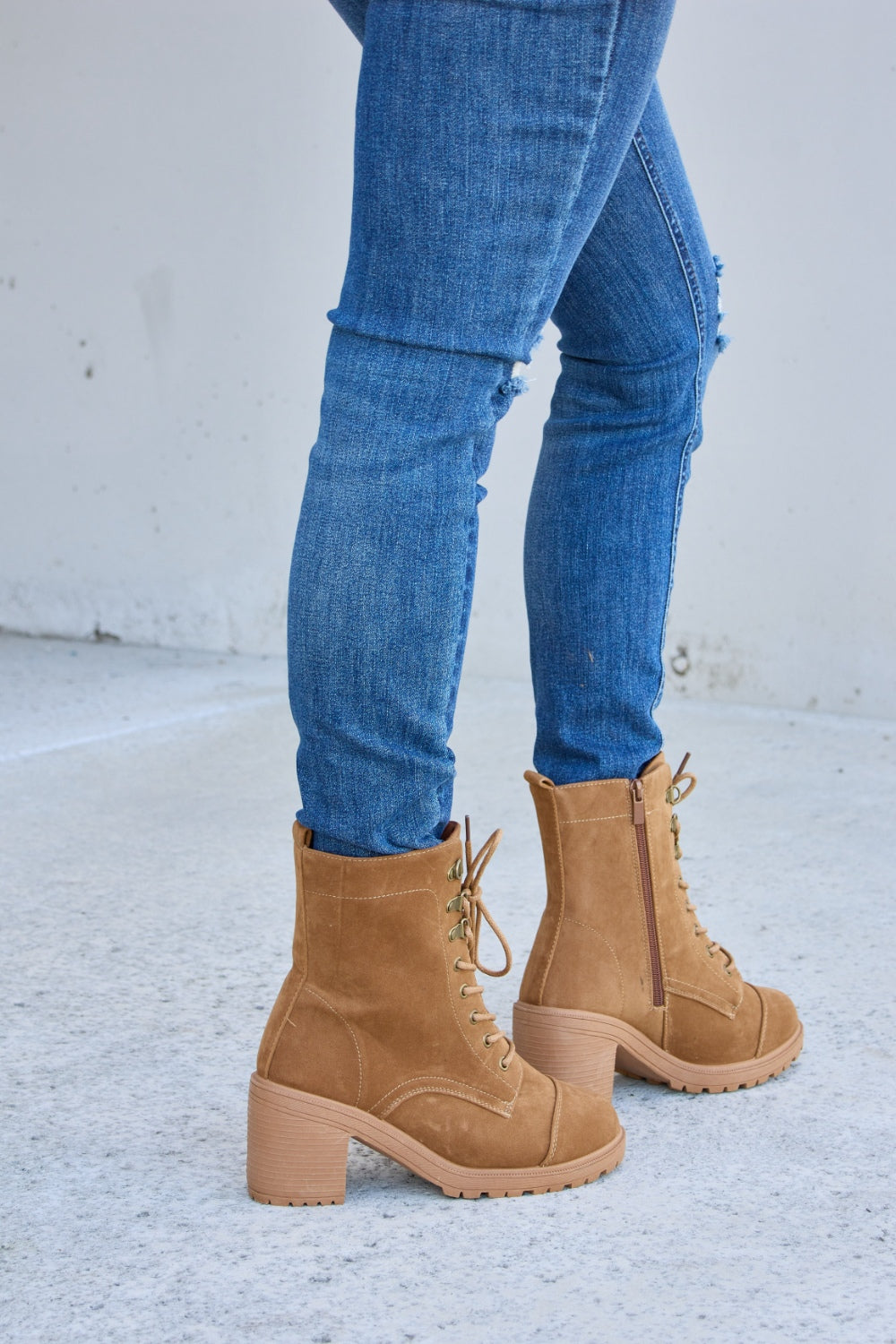 A person wearing distressed blue jeans and Forever Link Lace-Up Zipper Detail Block Heel Boots stands on a concrete surface.