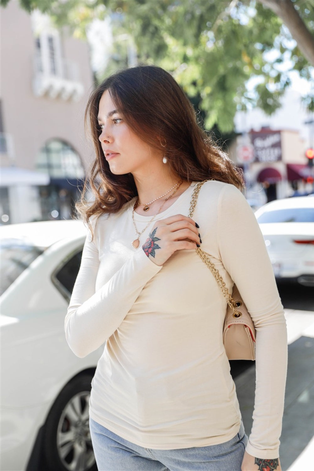 A woman with long hair stands outdoors in a classic wardrobe staple: the Stella Cruz Round Neck Long Sleeve T-Shirt in beige paired with jeans, while holding a purse. A building and car are visible in the background.