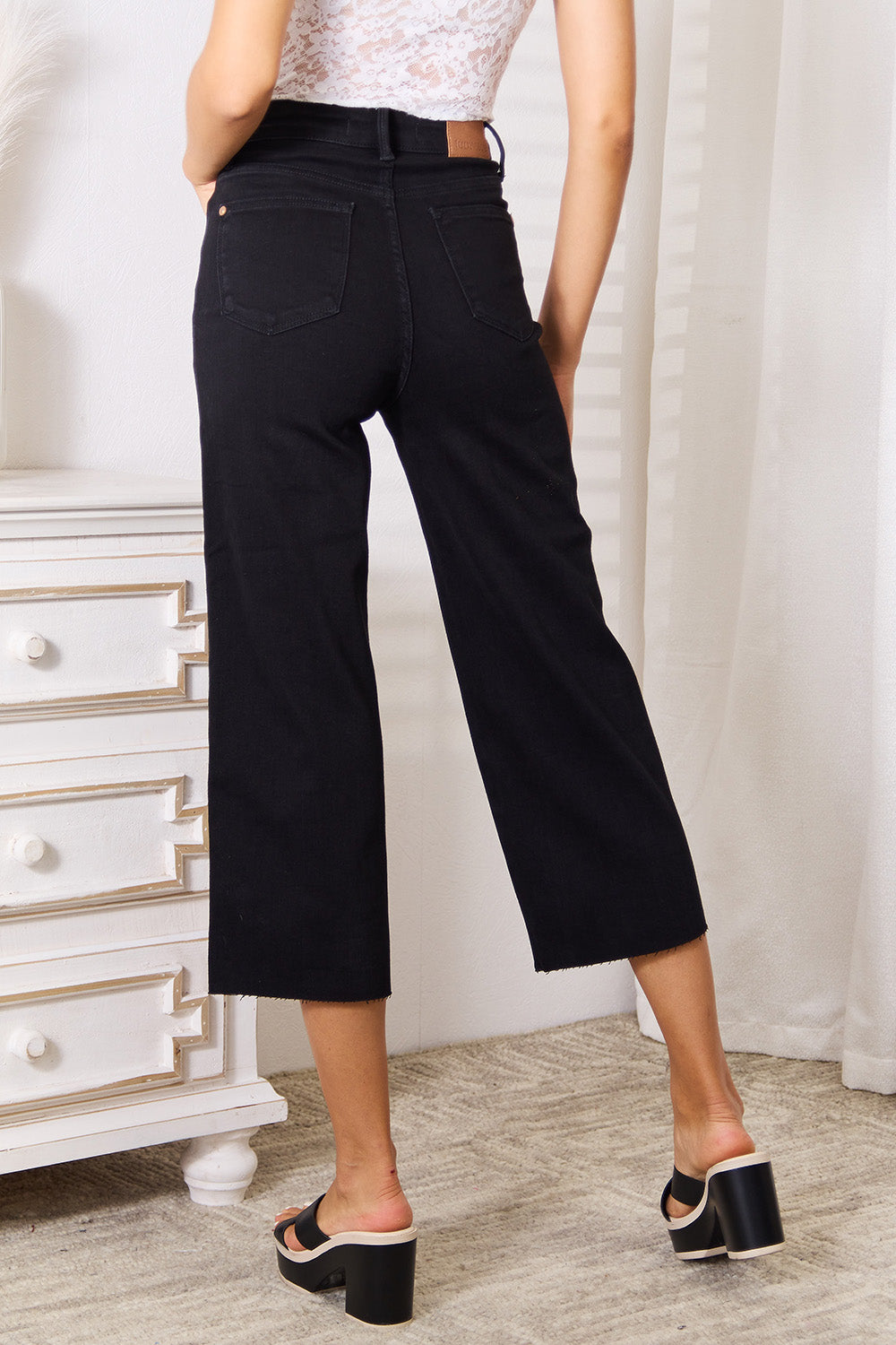 A person wearing Judy Blue Full Size High Waist Wide Raw Hem Cropped Jeans in black and a white lace top stands next to a white dresser adorned with a vase and candle.