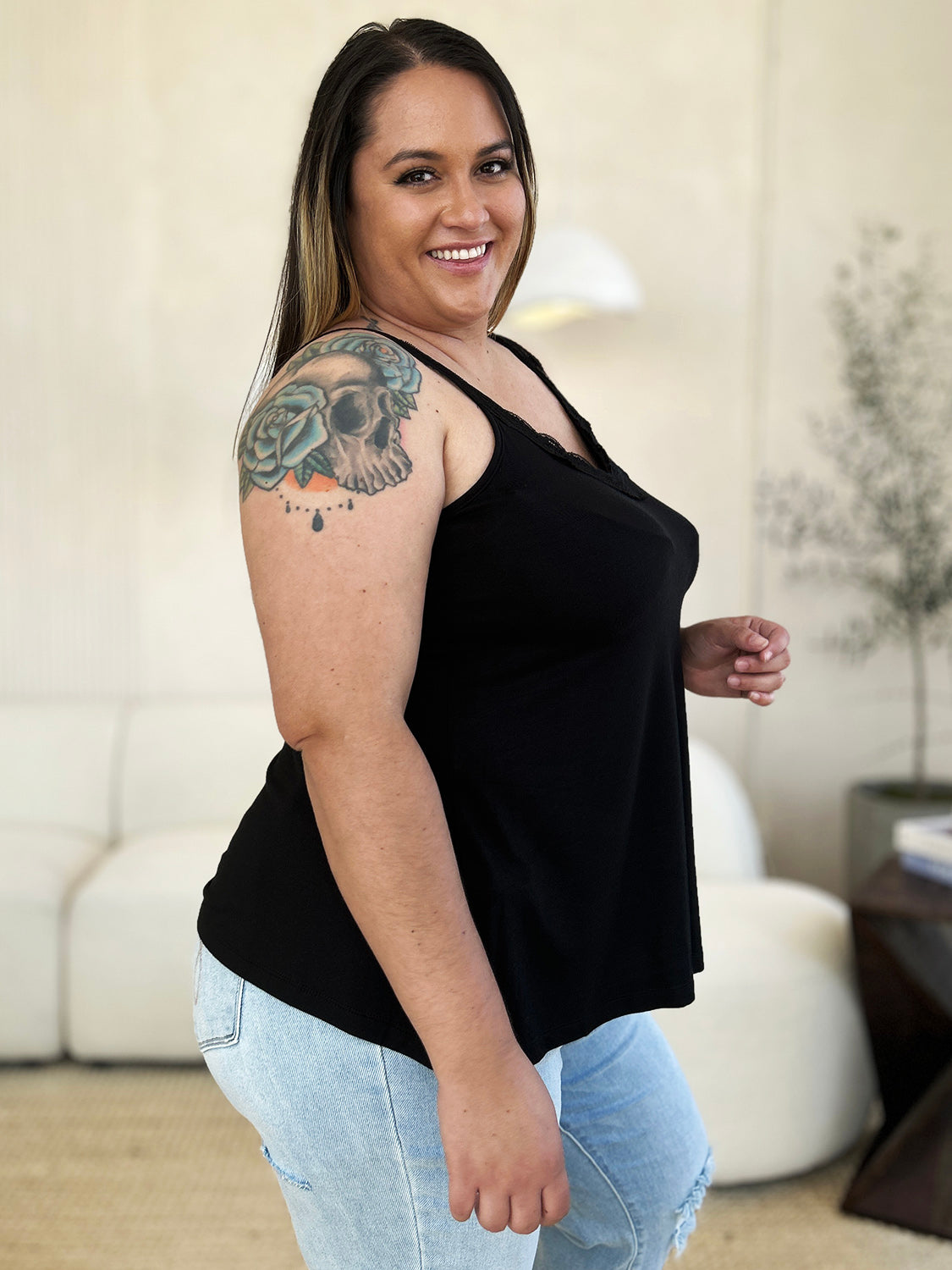 A woman with long hair wearing sunglasses, a slightly stretchy pink "Basic Bae Full Size Lace Detail V-Neck Cutout Cami," and basic style blue jeans stands against a concrete wall, looking to the side with one hand touching her upper chest.