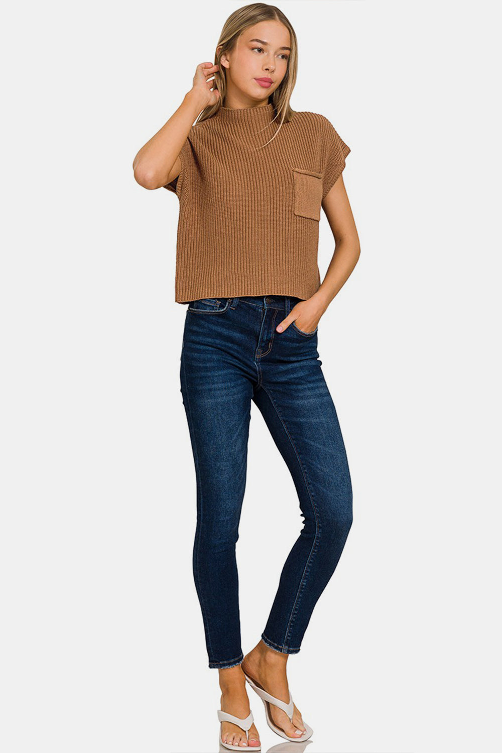 A person with long hair is wearing a Zenana Mock Neck Short Sleeve Cropped Sweater in brown and blue jeans. They are touching their hair with one hand and looking to the side, showcasing the casual comfort of their attire.