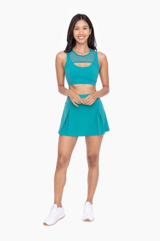 A person wearing a Two Pleat Active Tennis Skort in turquoise, featuring four-way stretch, and white sneakers stands against a plain background.