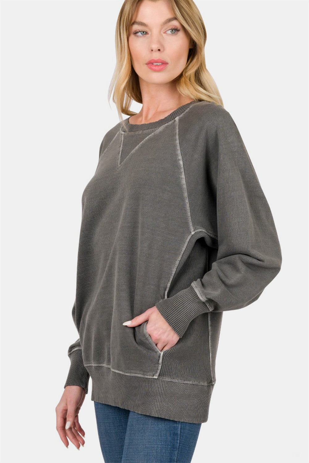 A person stands wearing a Zenana Round Neck Long Sleeve Sweatshirt, featuring a loose fit, dark grey color with front pockets and light stitching, paired with blue jeans. The 100% cotton fabric ensures comfort and durability.