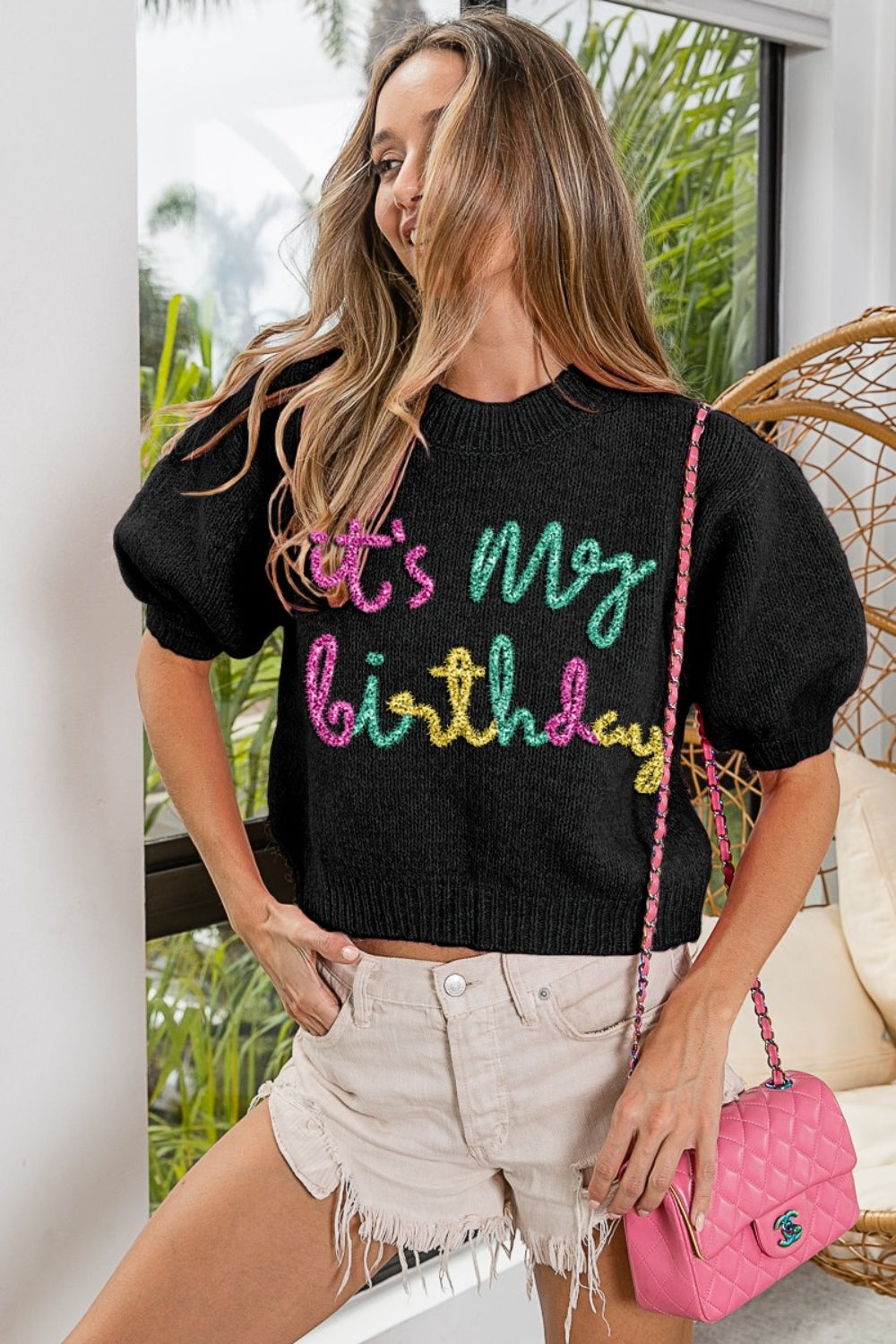 A woman poses against a pink wall, wearing a BiBi Metallic Letter Puff Sleeve Hairy Sweater that reads "It's My Birthday," beige shorts, and novelty glasses with birthday candles.