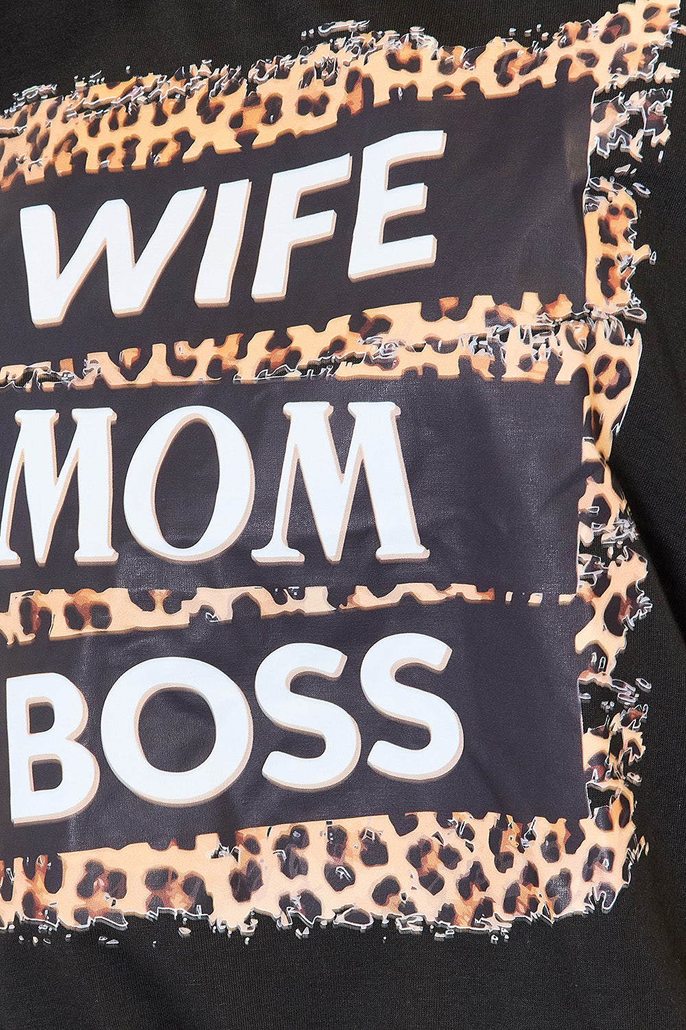 A woman beams with confidence as she dons a Simply Love WIFE MOM BOSS Leopard Graphic T-Shirt, the bold wording displayed prominently across the front. Beside a white dresser, she exudes empowerment and grace, embodying strength in her daily life.