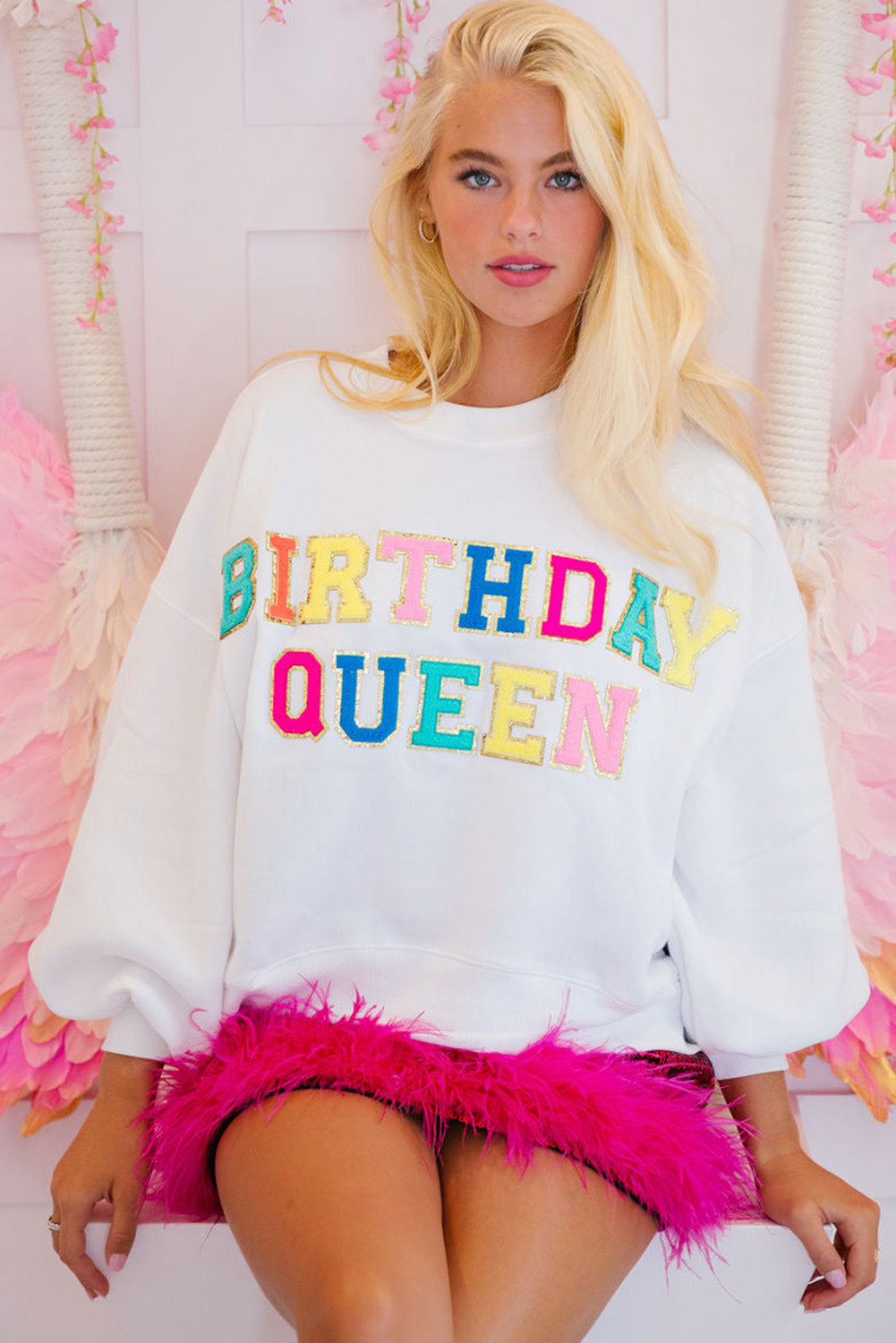 A person with long blonde hair is wearing a White BIRTHDAY QUEEN Graphic Balloon Sleeve Sweatshirt, paired with a pink feathered skirt, and sitting against a beautifully decorated backdrop.