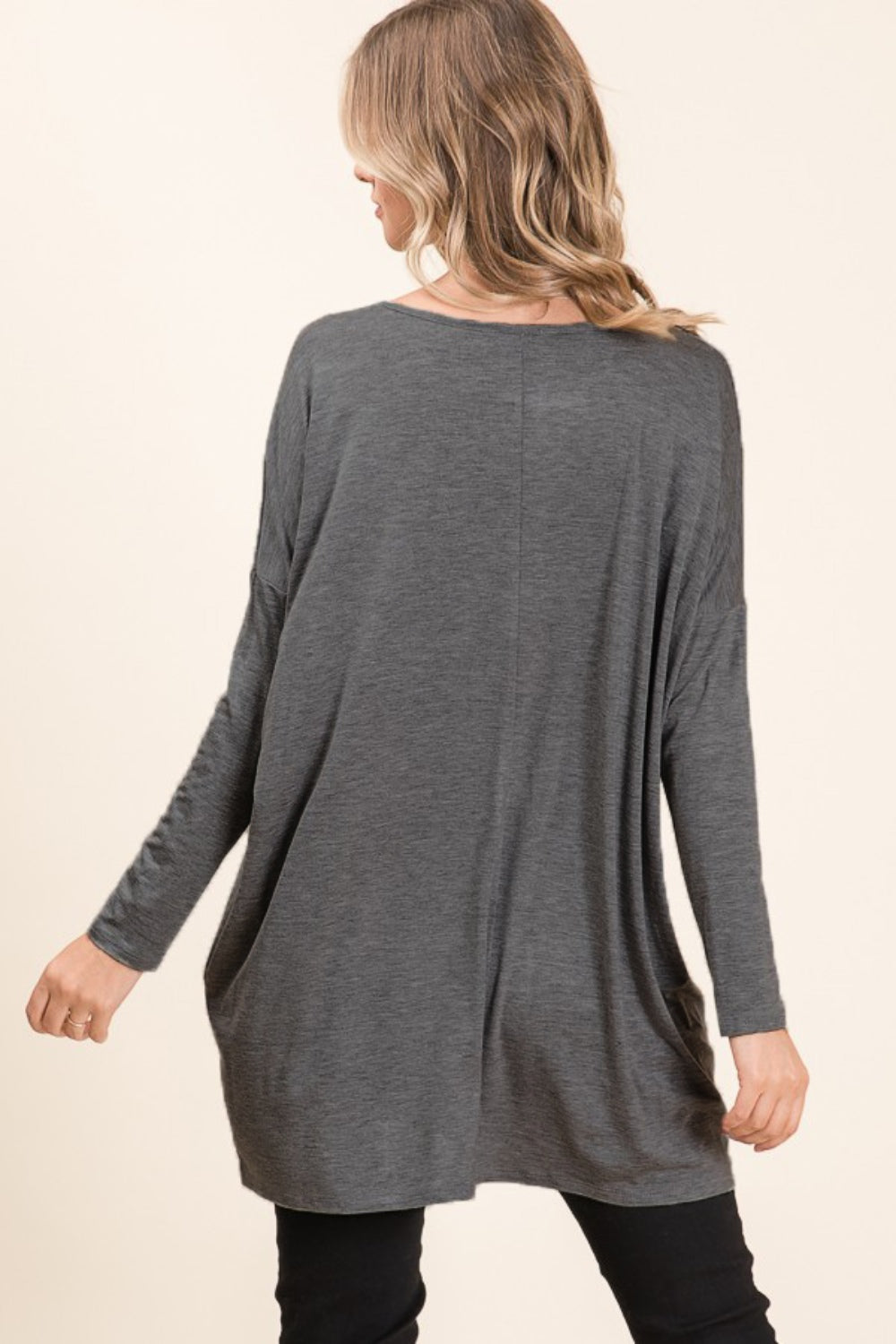 A woman with long hair is wearing a cozy, BOMBOM Round Neck Long Sleeve Oversized Top and blue jeans. She is smiling with one hand touching her head and the other in her pocket. Her comfortable chic wardrobe includes a long necklace, adding a stylish touch to her relaxed outfit.