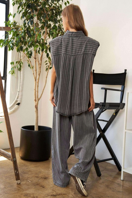 A person wears a casual outfit called the Stripe Button Down Shirt and Long Pants Set, which features a loose-fitting pinstriped design, paired with sneakers. They stand indoors near a potted plant and a director's chair.