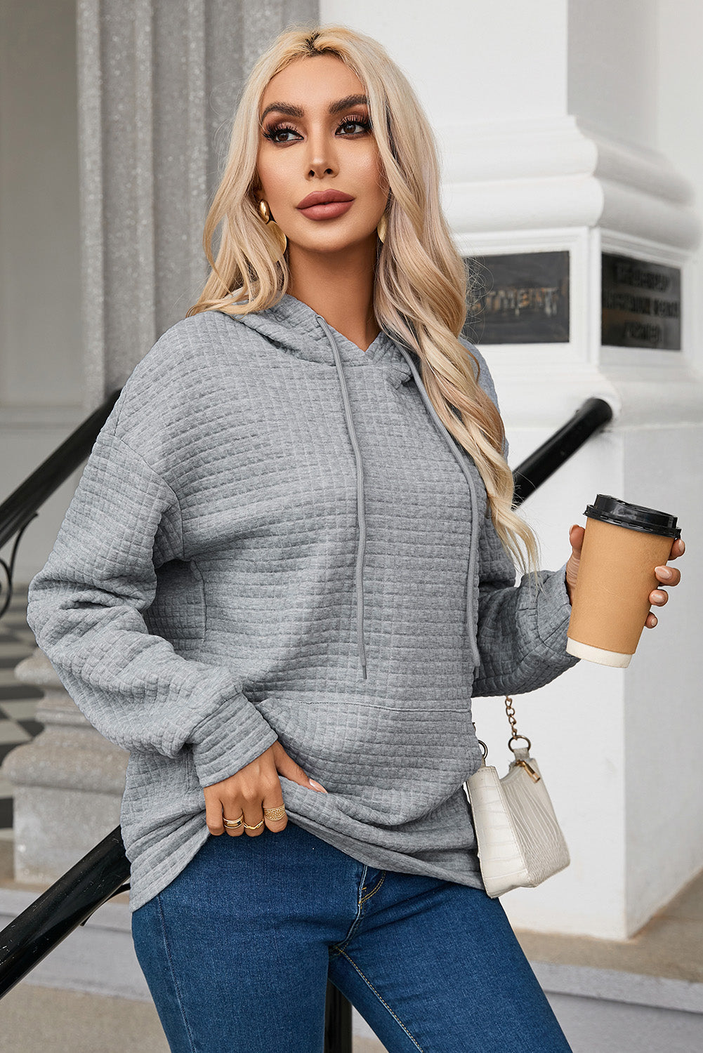 A person with long blonde hair is seen from behind, wearing a Gray Quilted Kangaroo Pocket Drawstring Hoodie and blue jeans. The relaxed fit of the textured fabric gives a casual vibe as they stand outdoors near a staircase with a metal railing.