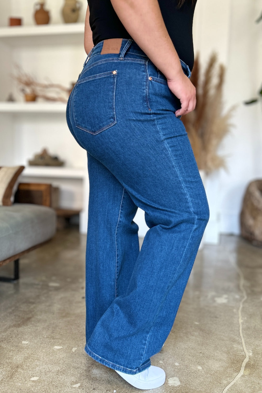 A person wearing Judy Blue Full Size High Rise Straight Jeans, a black top, and white shoes stands gracefully on a polished concrete floor.