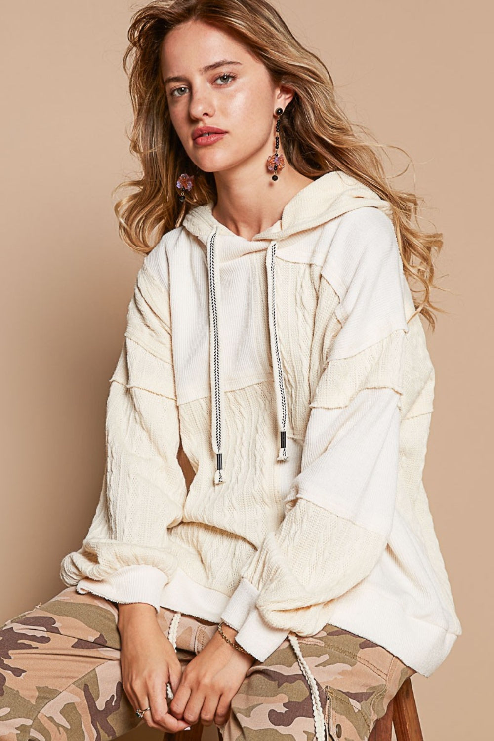 A person wearing a POL Exposed Seam Hooded Knit Top and camouflage pants sits on a stool against a beige background.