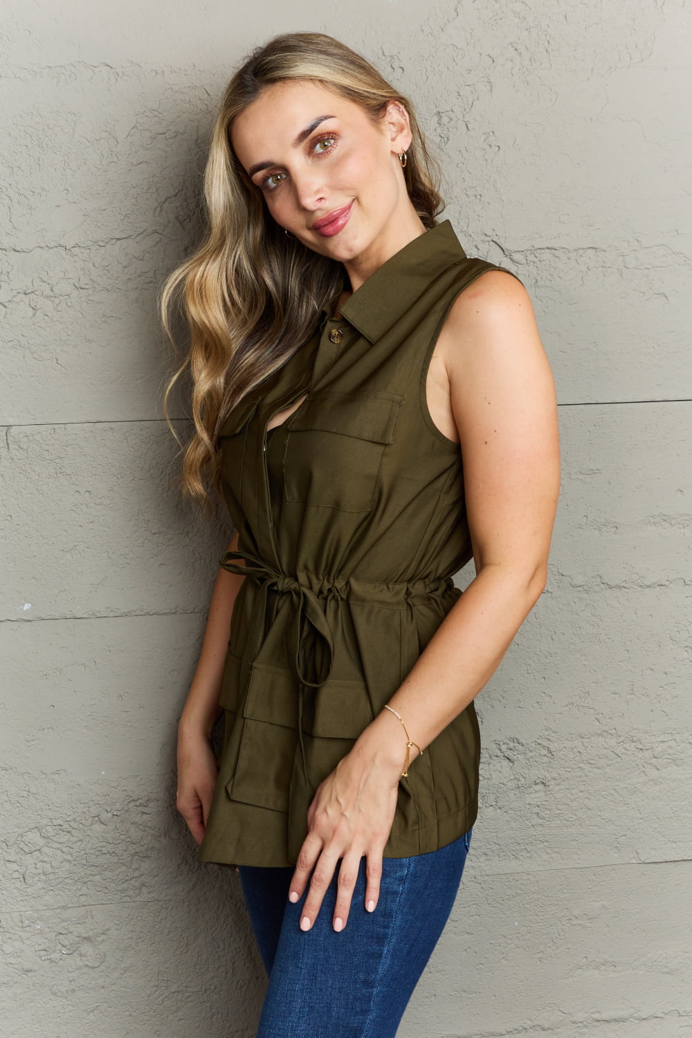 A woman with long, wavy hair wearing the Ninexis Follow The Light Sleeveless Collared Button Down Top in olive-green high-quality fabric and a belted waist stands against a textured gray wall, smiling.