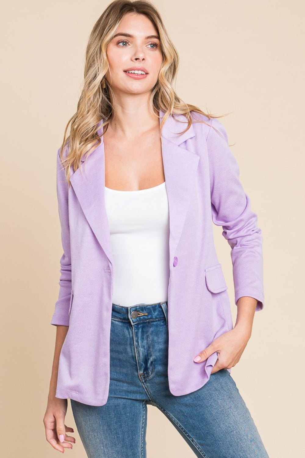 A person with long, wavy hair is wearing the Culture Code Button Up Long Sleeve Slit Blazer in light purple over a white top and blue jeans, standing against a beige background.