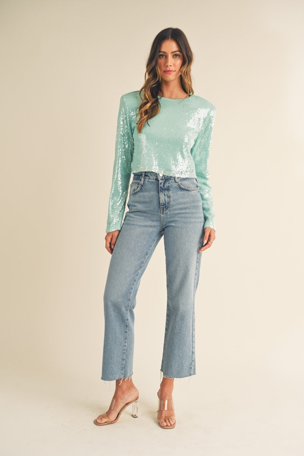 A person with long hair, wearing a fashion-forward, seafoam green MABLE Shoulder Padded Sequin Crop Top and high-waisted blue jeans, stands against a plain background.