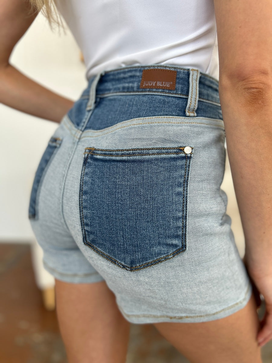 A person models indoors beside a beige chair and a plant, sporting a white t-shirt with the Judy Blue Full Size Color Block Denim Shorts for an effortlessly bold, eye-catching look.