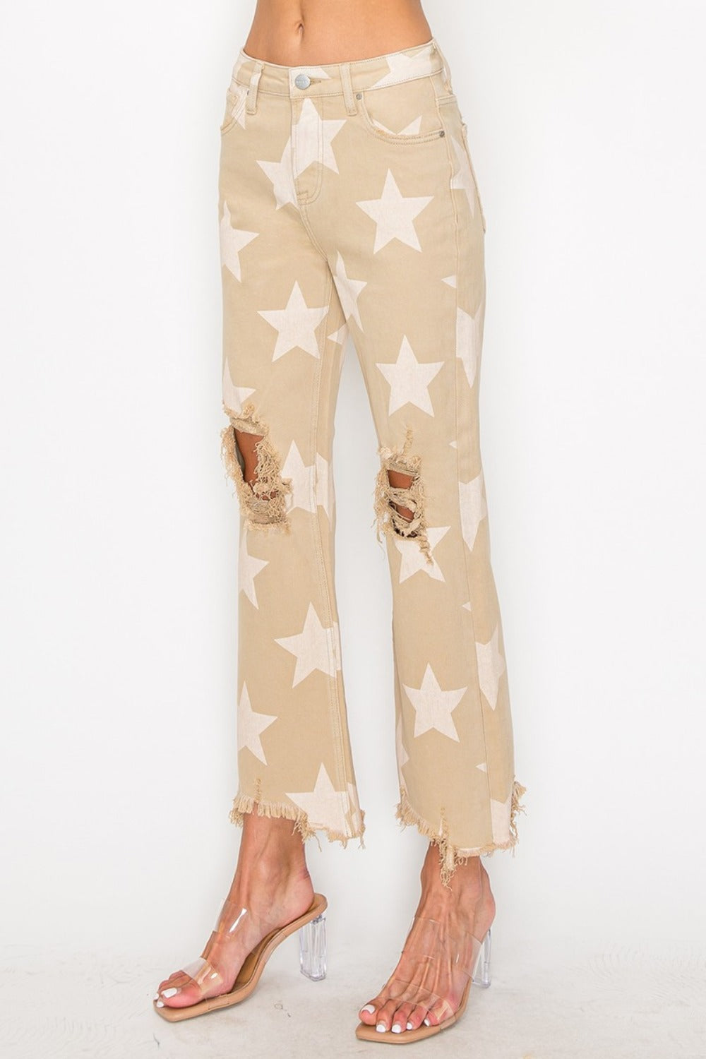 Someone dressed in RISEN Full Size High Rise Star Printed Frayed Hem Jeans with ripped knees, complemented by clear heels.