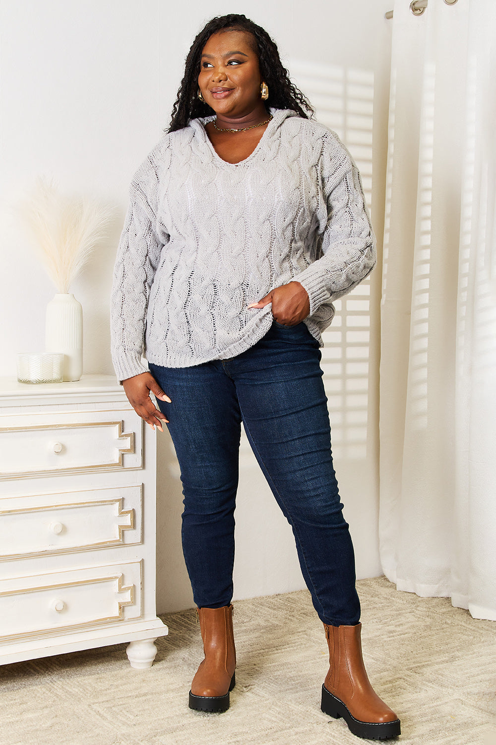 A person with long hair wearing a cozy Woven Right Cable-Knit Hooded Sweater and dark jeans is standing indoors looking to the side.