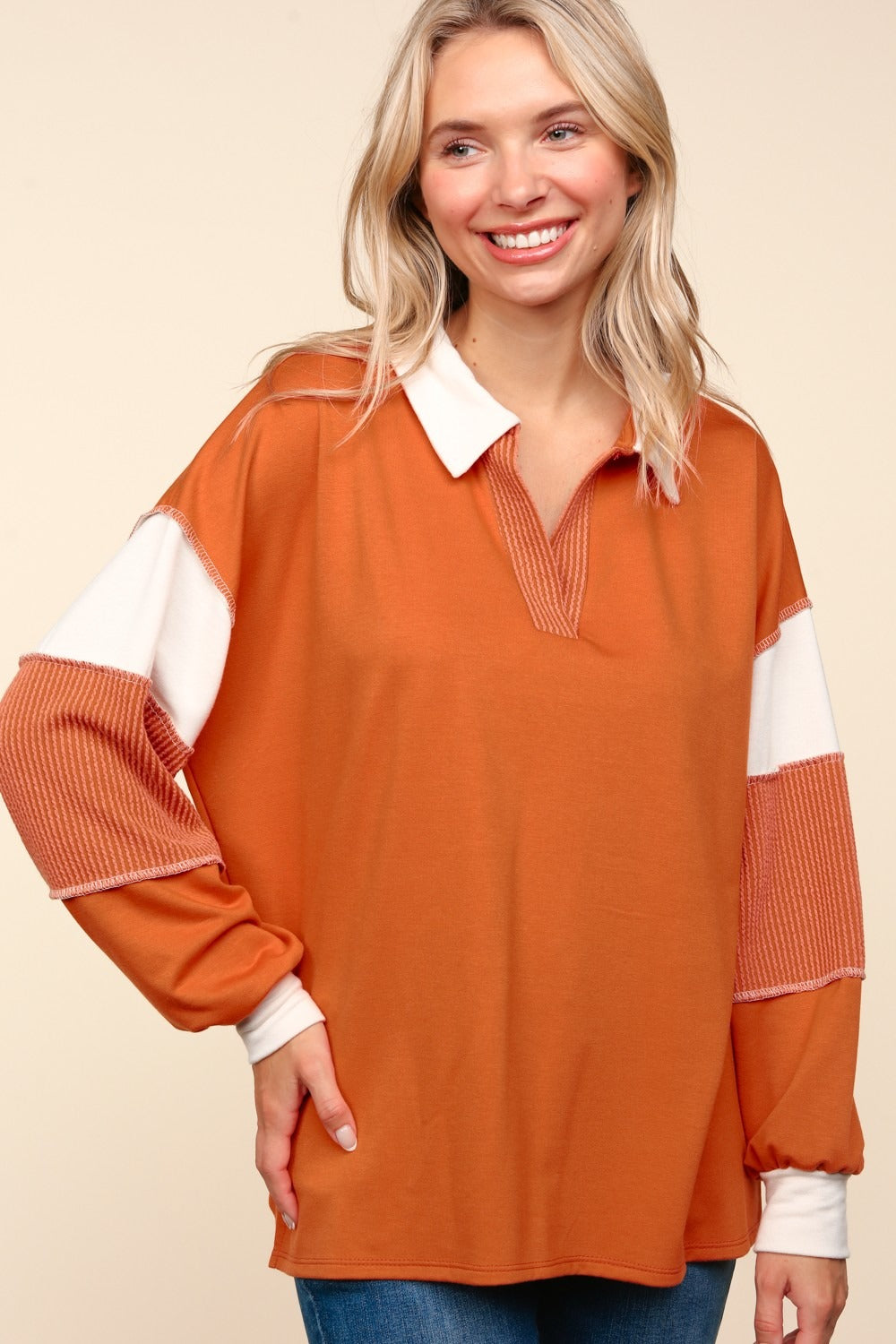 A person with blonde hair is wearing the Haptics Color Block Exposed Seam Long Sleeve Top in orange with white and orange sleeves, paired with blue jeans, standing against a beige background.