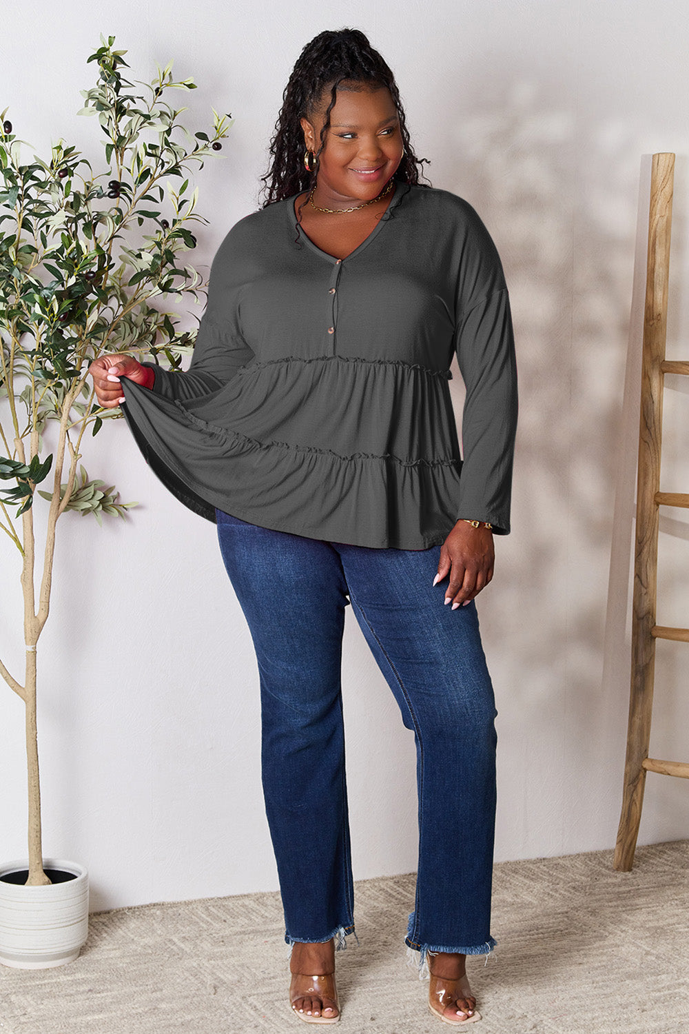 Person stands indoors near a plant, wearing the Double Take Half Button Long Sleeve Ruffle Hem Blouse in gray and blue jeans, smiling while looking to the side. The slightly stretchy fabric of the blouse adds comfort while its buttoned design offers a touch of elegance.