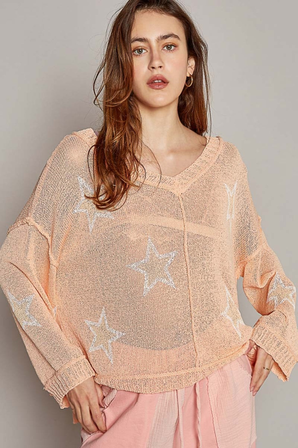 A woman with long hair is sitting down, wearing a fashionable POL V-Neck Long Sleeve Star Print Knit Top in a peach, loose-knit design. The top is paired perfectly with matching peach pants. She looks directly at the camera with a neutral expression.