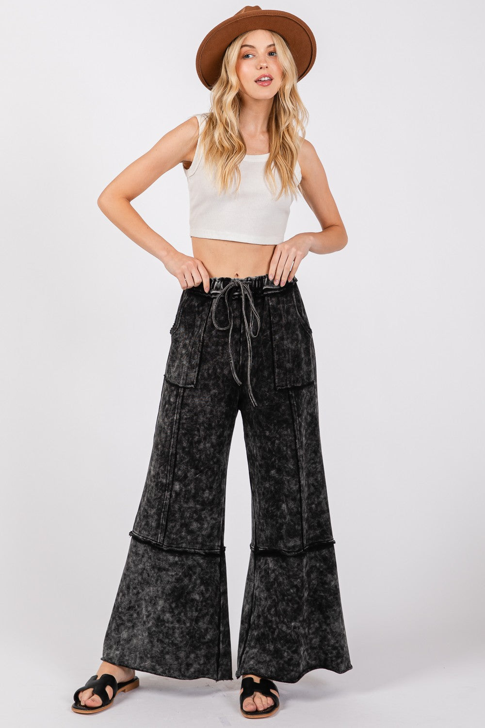 A woman stands against a plain background wearing a brown hat, white crop top, SAGE + FIG Mineral Washed Terry Wide Leg Pants with an elastic waistband and drawstring, and black sandals. Her hands rest on her waist.