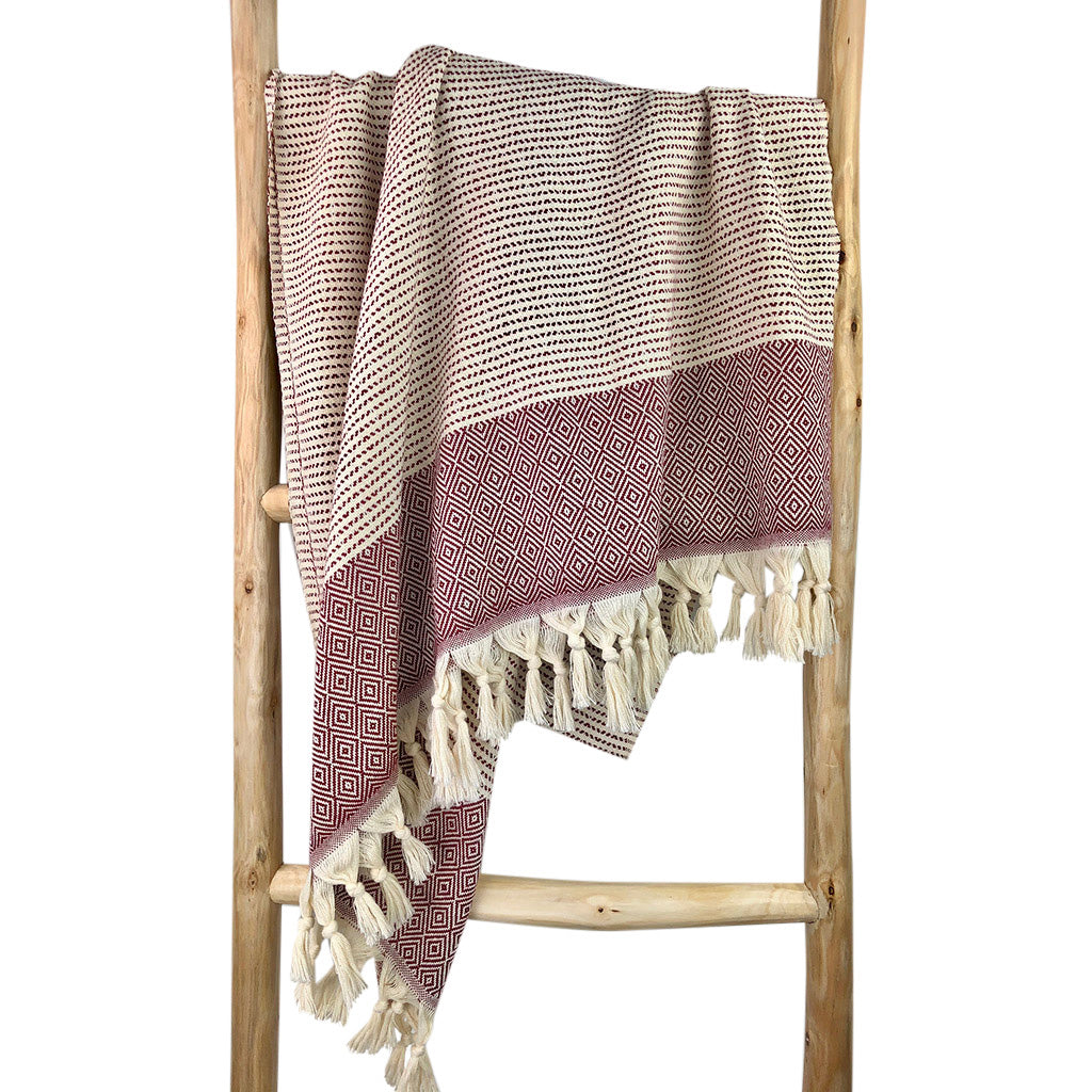 The Diamond Stripe Turkish Towel is crafted from luxurious Turkish cotton and features a folded, patterned design with a black and white striped top section and a red geometric-patterned bottom, complete with tasseled fringe at one end. It's perfect for adding elegance to beach outings or home decor.