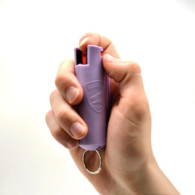 Guard Dog Pepper Spray keychain with integrated remote on a wooden table near a folded umbrella and a smartphone displaying text.