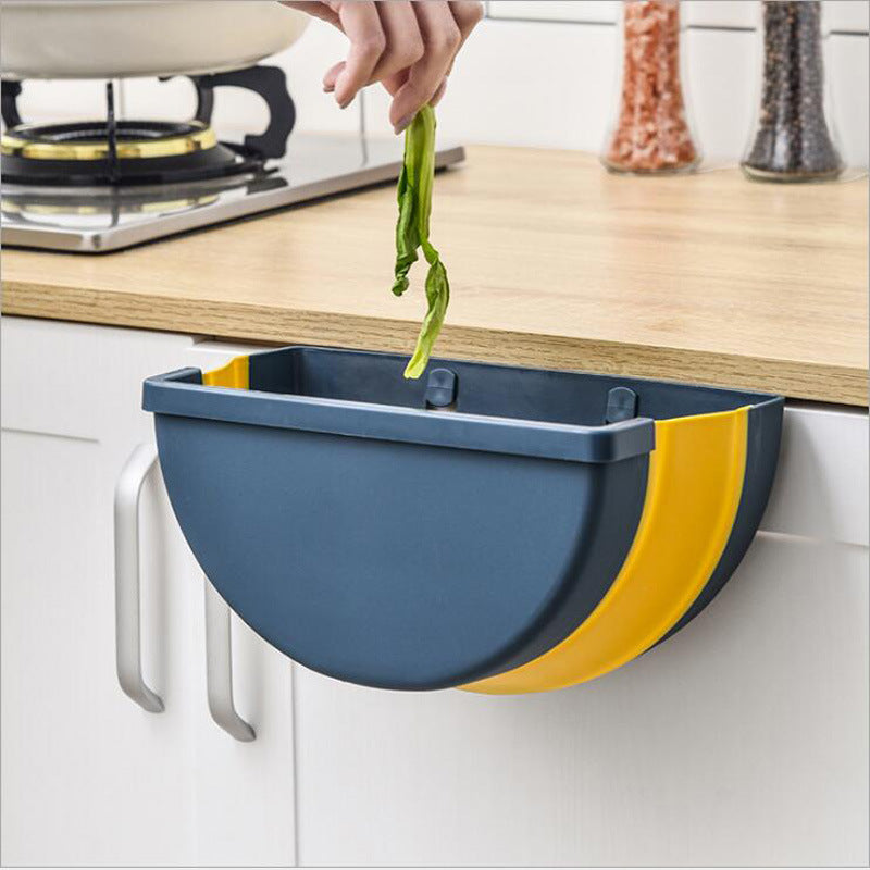 A hand is shown disposing of vegetable scraps into the Foldable Waste Bin, a blue and yellow semi-circular hanging trash can, attached to the edge of a kitchen counter. A stove and spices are visible in the background.