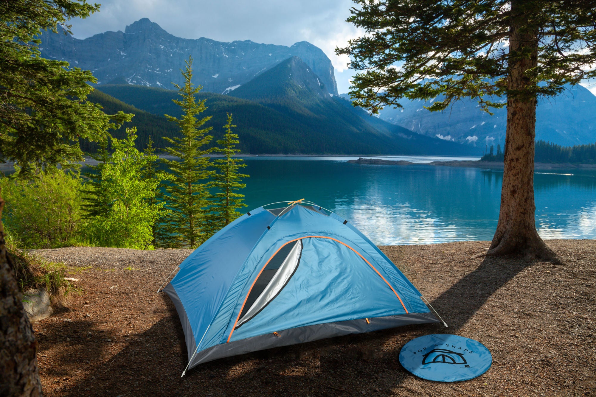 A blue POP-A-SHADE BLUE pitched by a serene lake surrounded by pine trees and towering mountains under a cloudy sky.