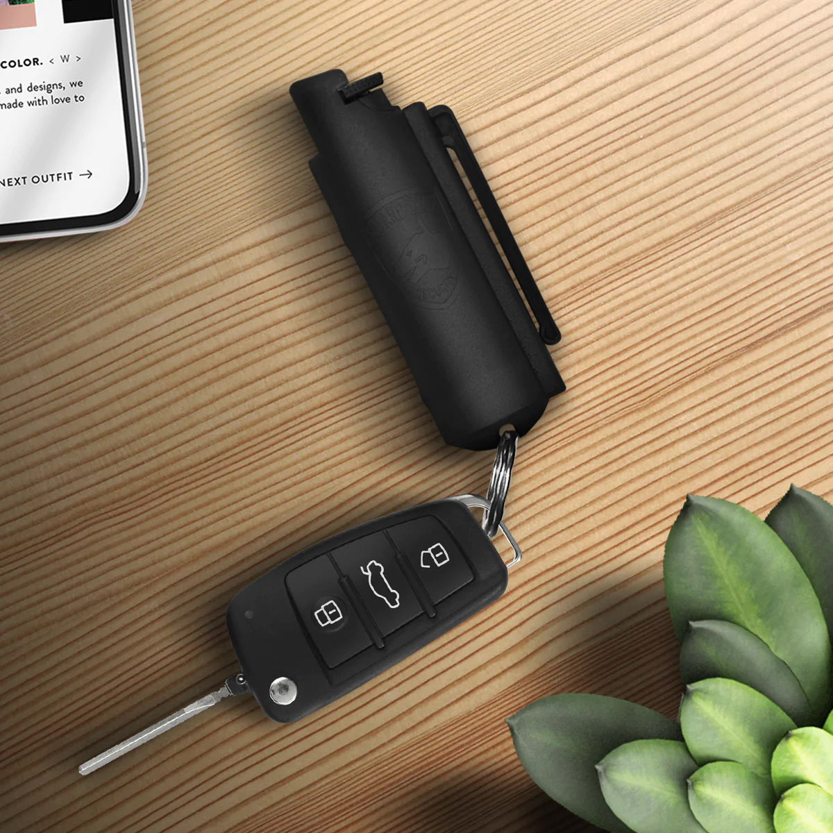 Guard Dog Pepper Spray keychain with integrated remote on a wooden table near a folded umbrella and a smartphone displaying text.