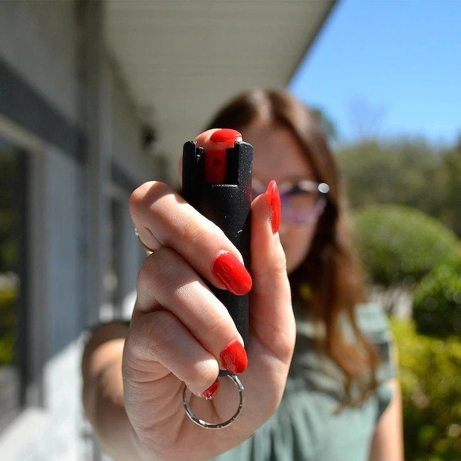 Guard Dog Pepper Spray keychain with integrated remote on a wooden table near a folded umbrella and a smartphone displaying text.