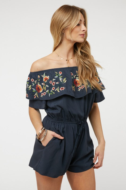 Wearing the stylish Floral Embroidered Off Shoulder Romper, a woman poses gracefully, showcasing its ruffle detail and making it an ideal addition to any summer wardrobe.