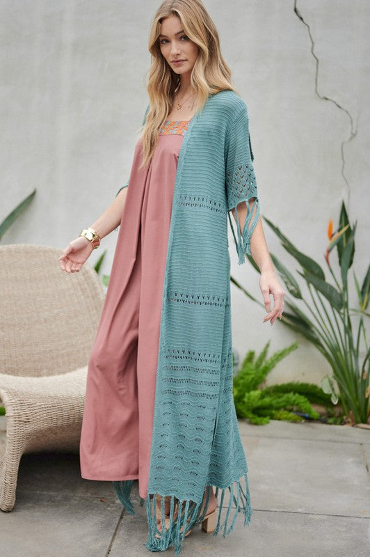 A woman in a green floral dress and a Solid Long Cardigan With Fringe exudes boho-chic vibes as she strolls on a patio, surrounded by outdoor furniture and lush plants.