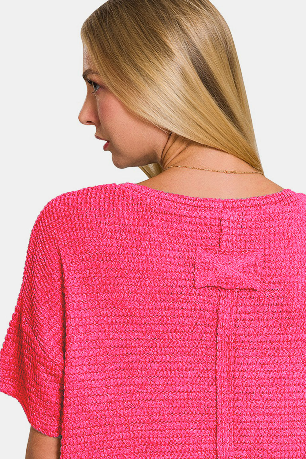 A woman wearing a loose-fitting, bright pink Zenana Drop Shoulder Short Sleeve Jacquard Knit Top with a V-neck and blue jeans touches her hair with one hand and looks at the camera.