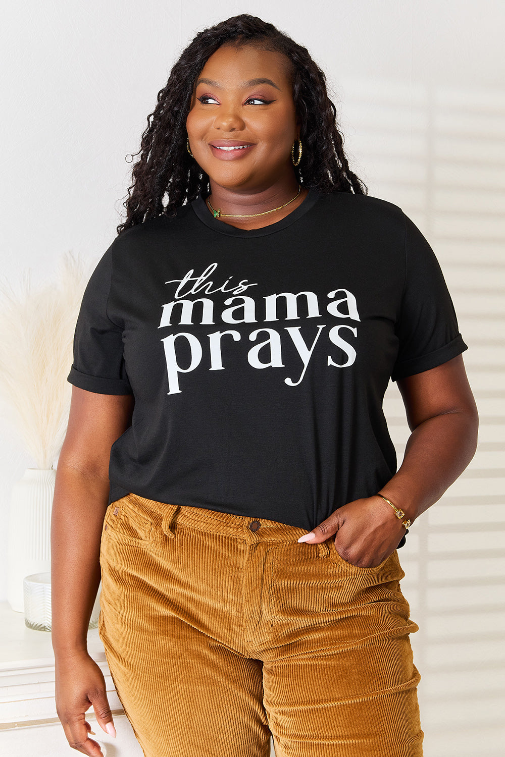 A woman wearing a Simply Love THIS MAMA PRAYS Graphic T-Shirt, paired with brown pants, stands gracefully in a softly lit room, exuding a sense of calm and spiritual gift.