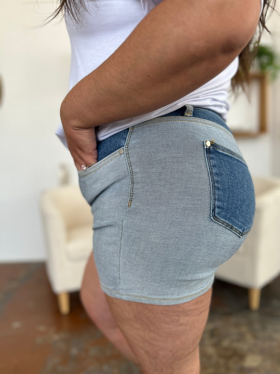 A person models indoors beside a beige chair and a plant, sporting a white t-shirt with the Judy Blue Full Size Color Block Denim Shorts for an effortlessly bold, eye-catching look.
