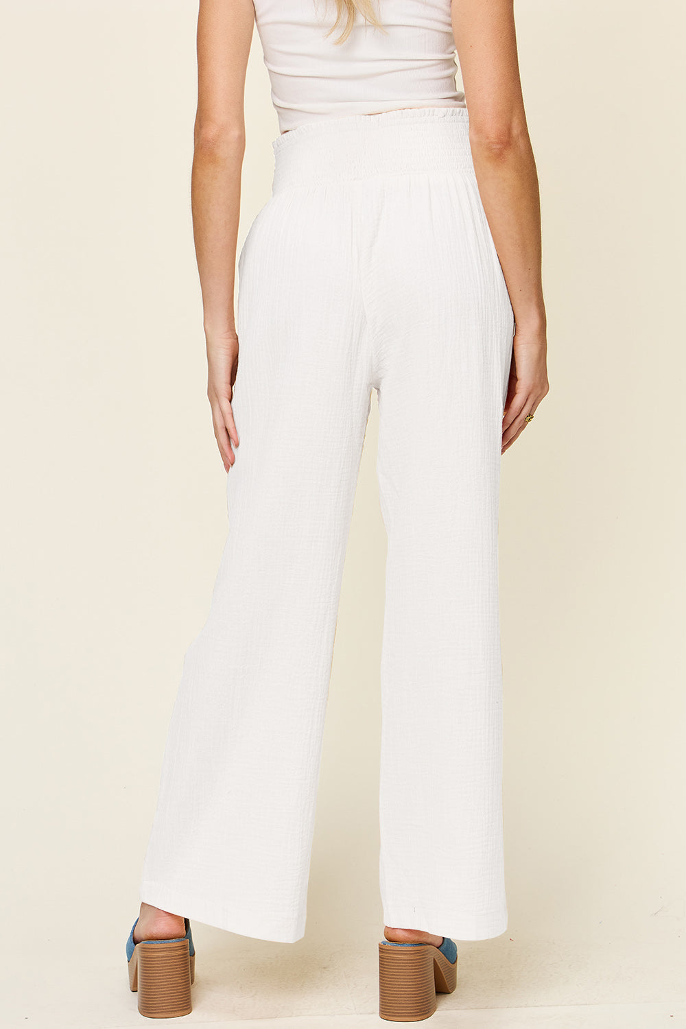 A person wearing Double Take Full Size Texture Smocked Waist Wide Leg Pants in black and a white 100% cotton top is standing with one hand on their hip. They are also wearing blue open-toe sandals.