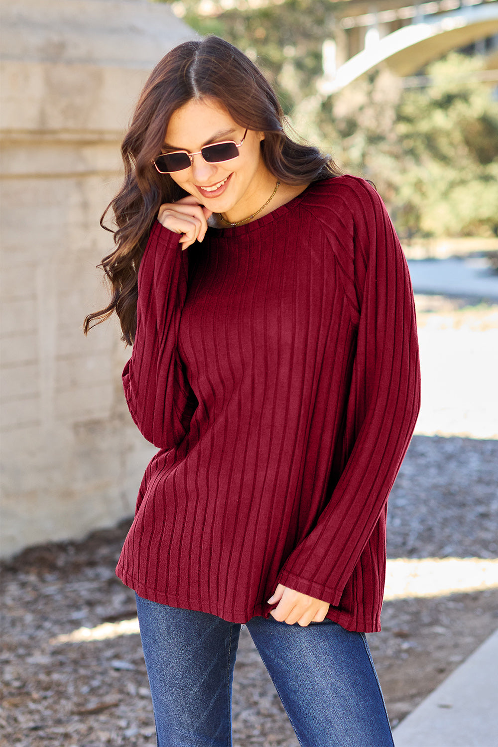 A woman with long brown hair wearing sunglasses and a Basic Bae full-size, slightly stretchy ribbed round neck long sleeve knit top in red, paired with blue jeans, is standing outdoors.