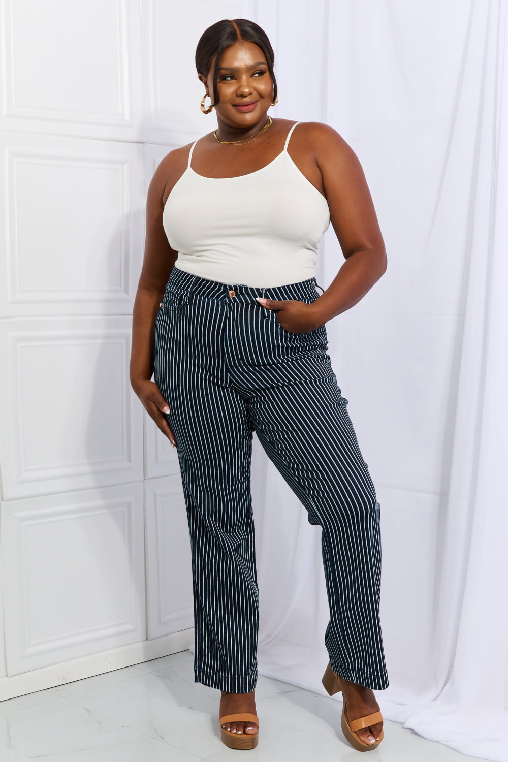 A person wearing Judy Blue Cassidy Full Size High Waisted Tummy Control Striped Straight Jeans, a white top, and tan platform sandals stands with one hand on their hip.